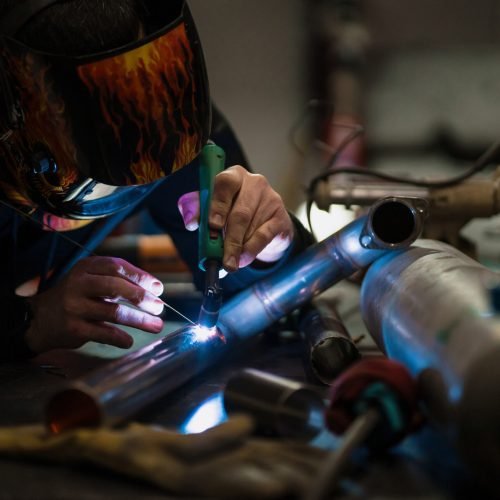 Worker in helmet with flames welding the steel part of a motorbike using an argon welding machine and gloves in a garage